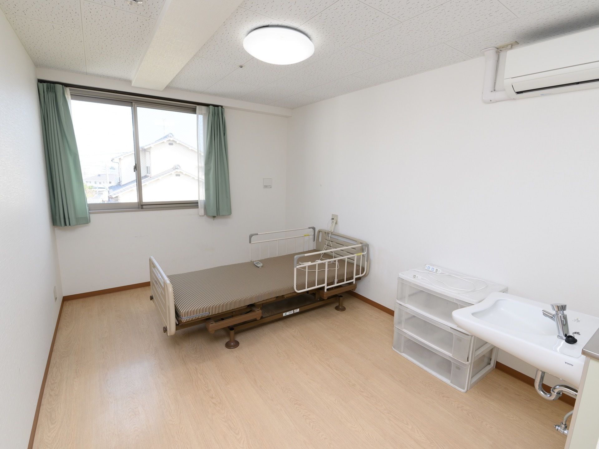 介護用ベッドや収納ケースの置かれた、１人用居室である。エアコン完備で手洗いコーナーもある。窓があり明るい。