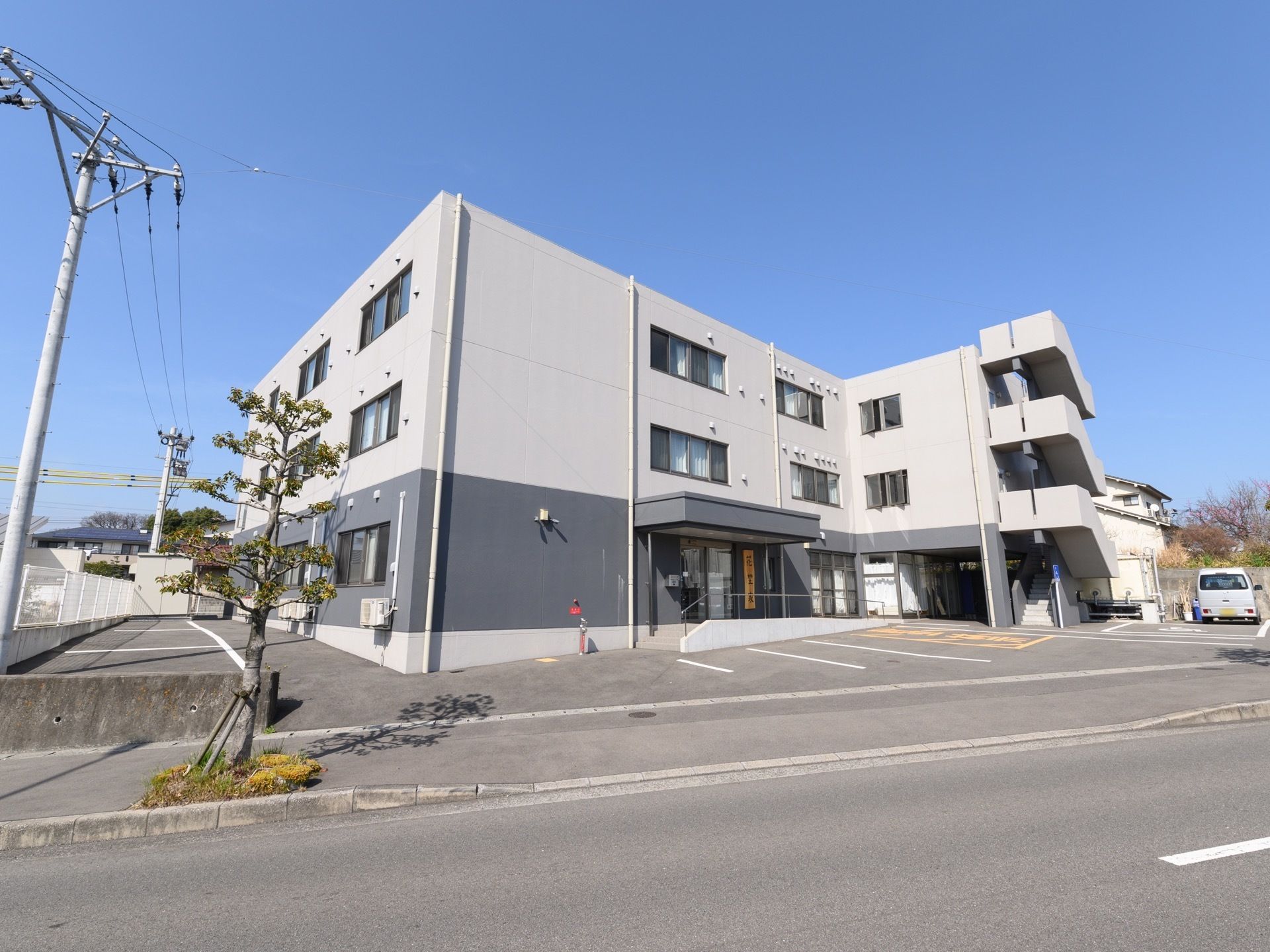 鉄骨造り３階建ての施設外観である。建物周囲は駐車スペースで、玄関前には車椅子専用駐車スペースとスロープがある。