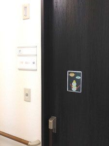 真っ黒い居室のドアにはフレームがあり、黄色いバケツをかぶった雪だるまのイラストが飾られている。居室のドアのそばまで手すりが付いている。