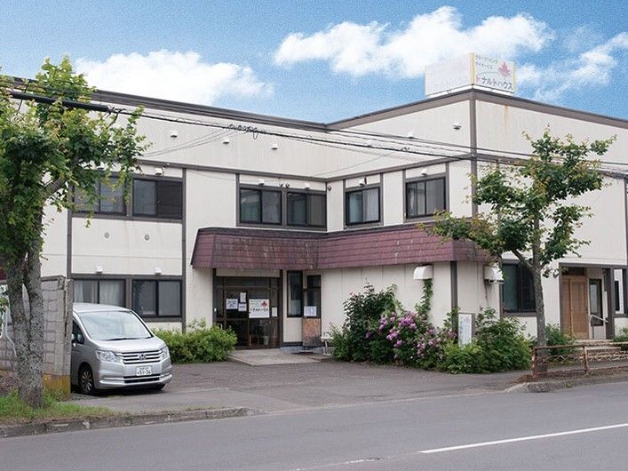 2階建ての建物は白い外壁で、エル字型、広い駐車場があり、草花が植えられ家庭的な雰囲気。