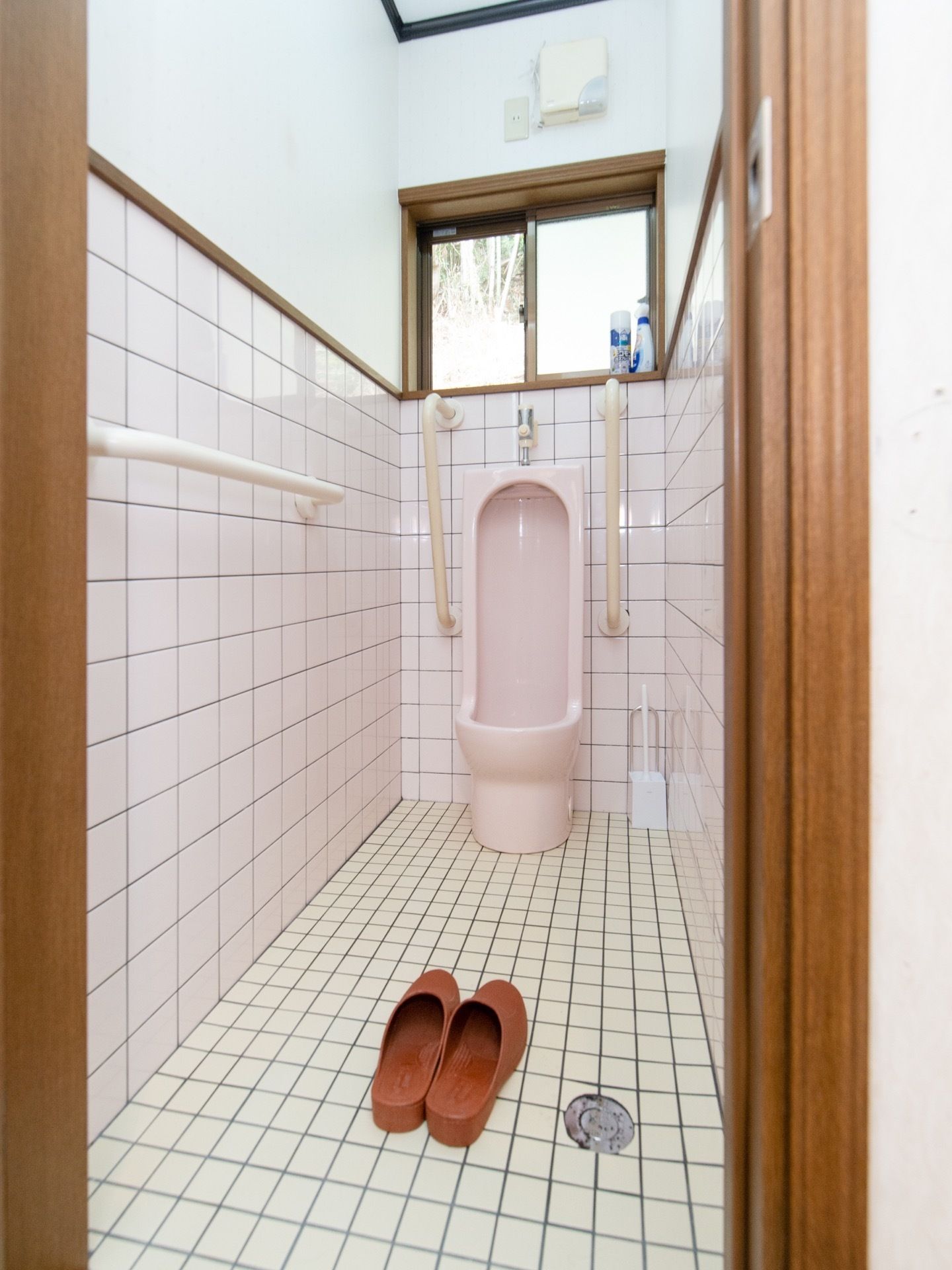 床と壁にタイルが貼られた男性用トイレがある。茶色いスリッパが並べて置いてあり、正面の小窓が開いている。