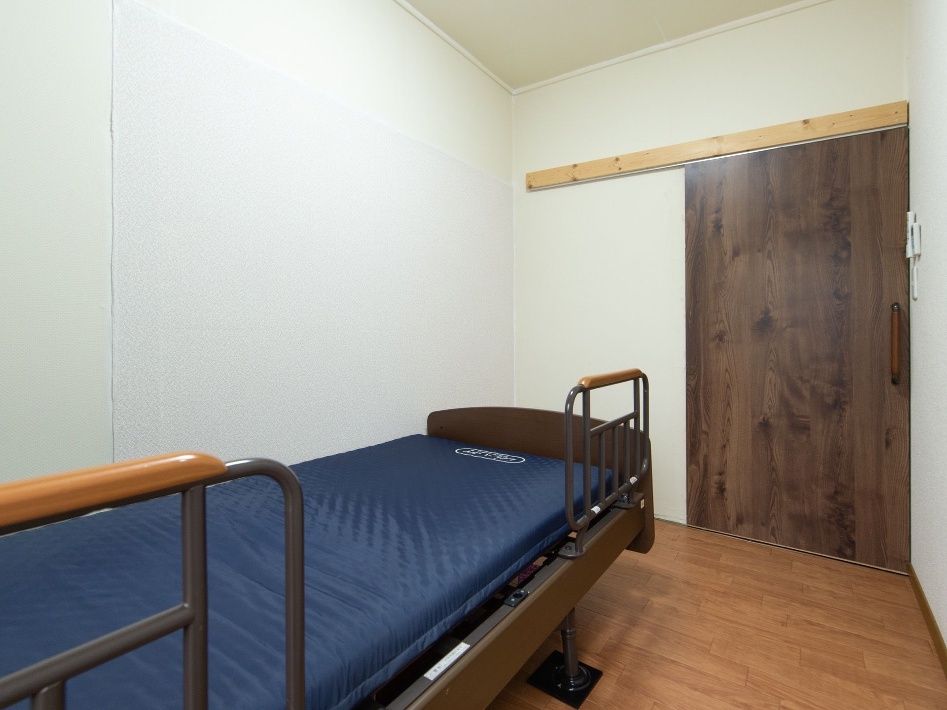 木目の大きなスライドドアの入り口がある。フローリングの床の上に青いマットで、手すりが設置されているベッドが置いてある。