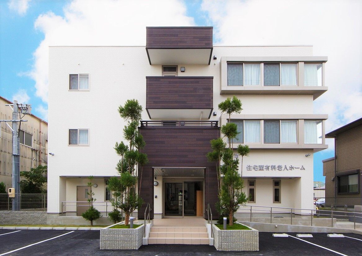 ベランダ付きの3階建ての建物。玄関には手すり付きのスロープがつき、白を基調とした明るい建物にはブラウンのベランダが付いている。