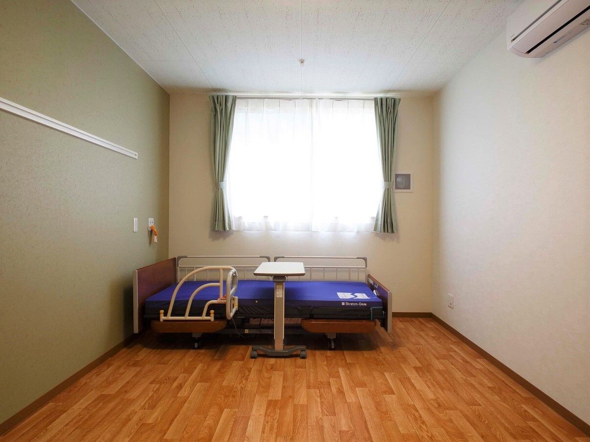 大きな窓が付いた明るいフローリングの居室には、ピクチャーレールやエアコン、介護用ベッドがあり、アクセントクロスが印象的。