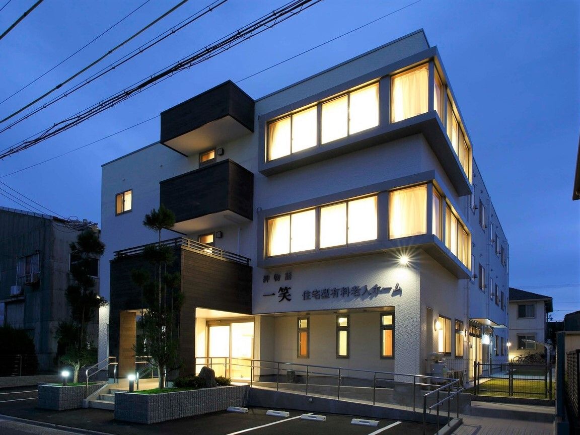 日が暮れてあたりが暗くなっている中、建物から漏れる明かりでシルエットが浮かび上がっている。3階建てでベランダがあり、駐車場も付いている。
