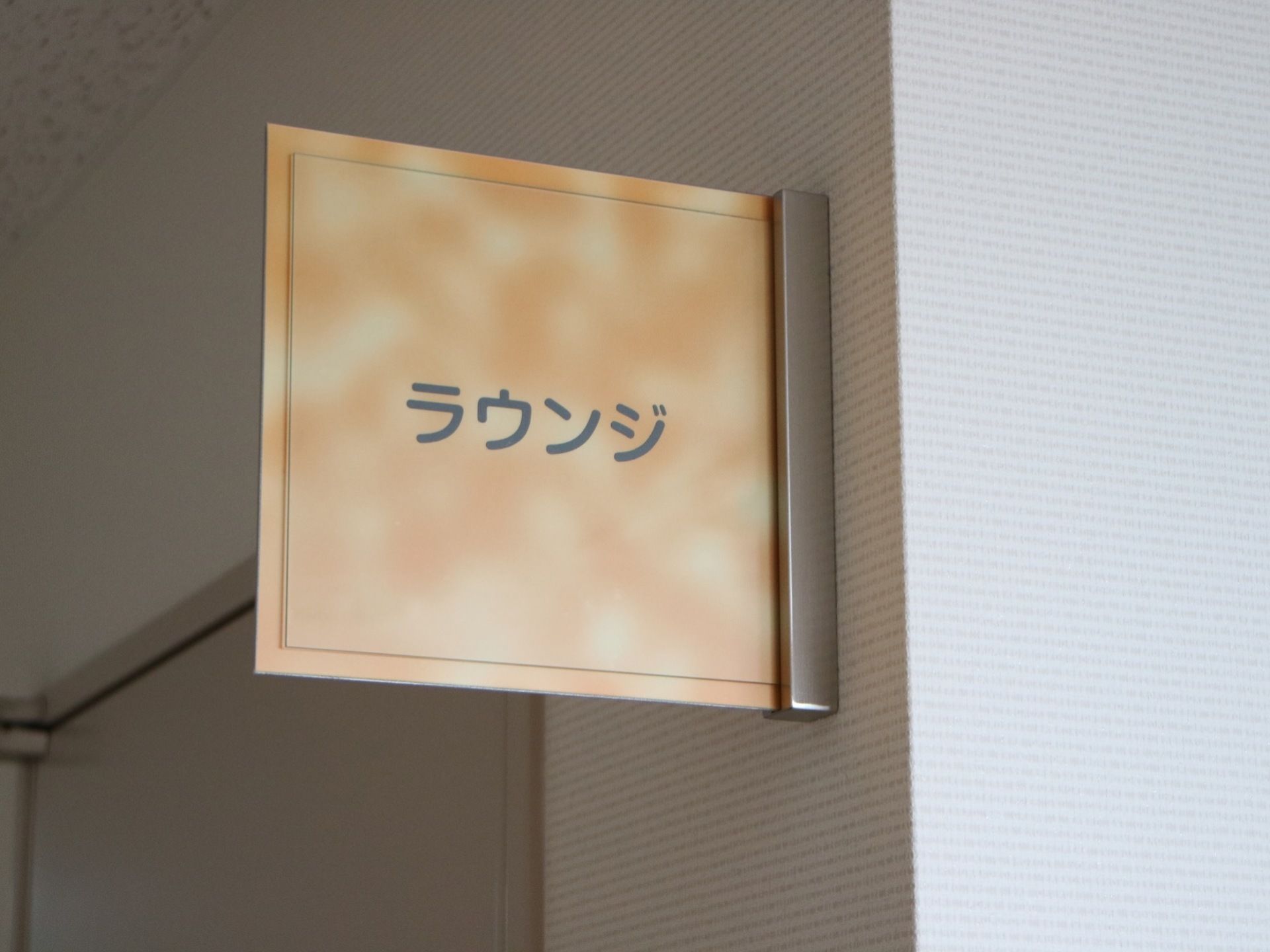 白い壁の廊下である。上部にラウンジと書かれたプレートが取り付けられている。奥に片開き式のドアが見える。