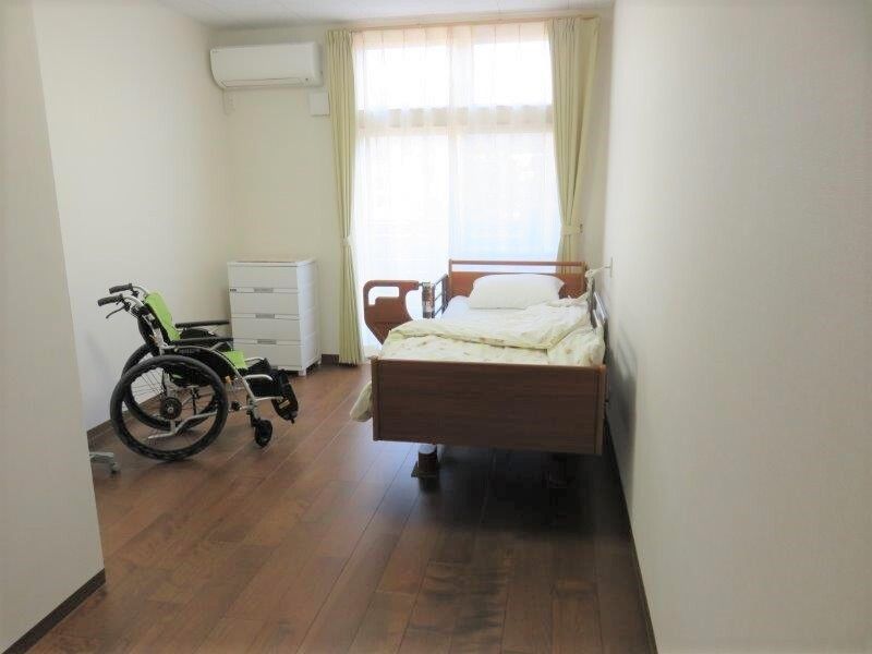 小さなワンルームタイプの部屋にベッドやタンス、車いすが置かれている様子