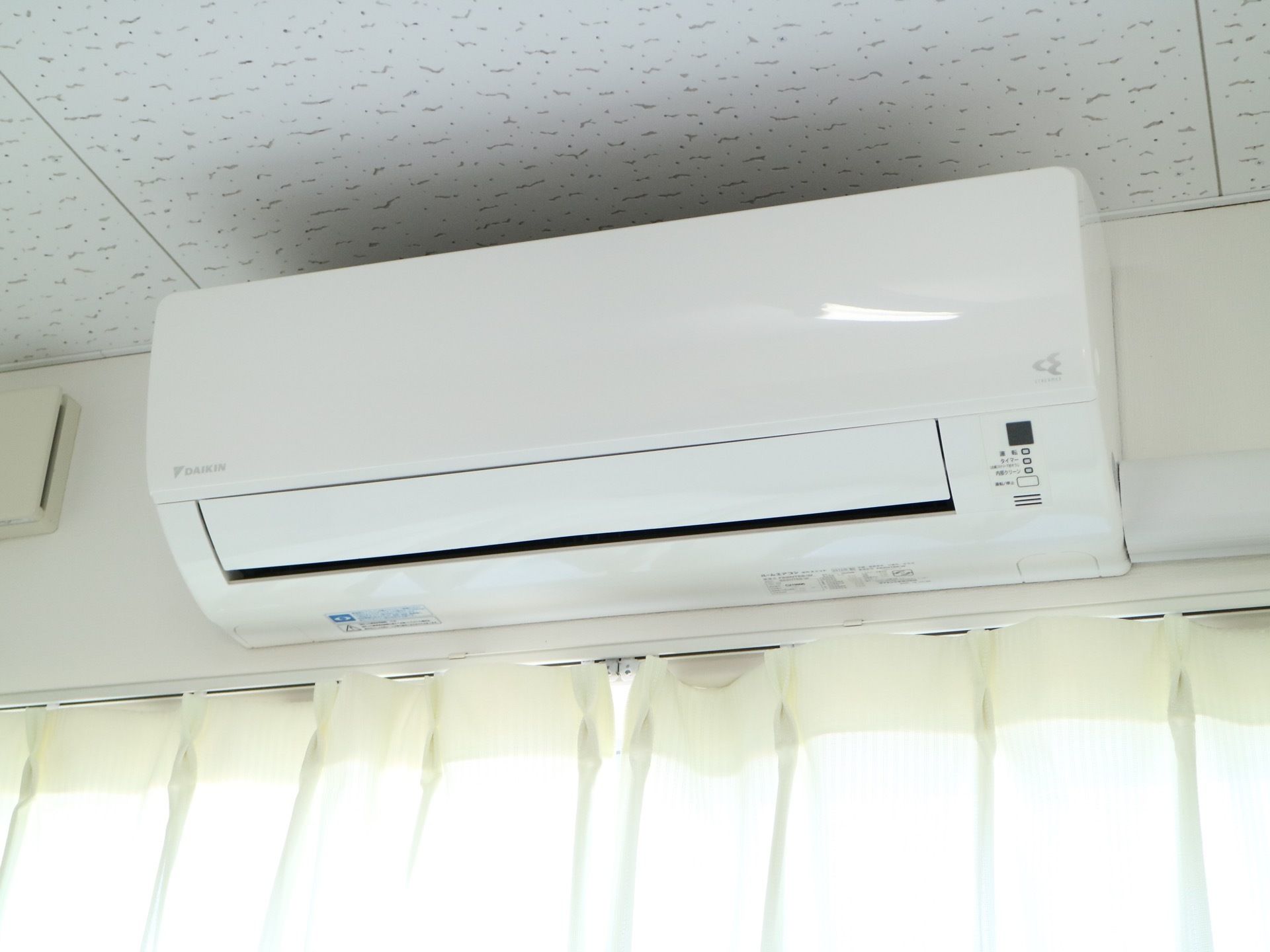 窓が備え付けられた居室である。上部には白色のエアコンが完備されている。天井は不規則な模様が施された、壁紙である。