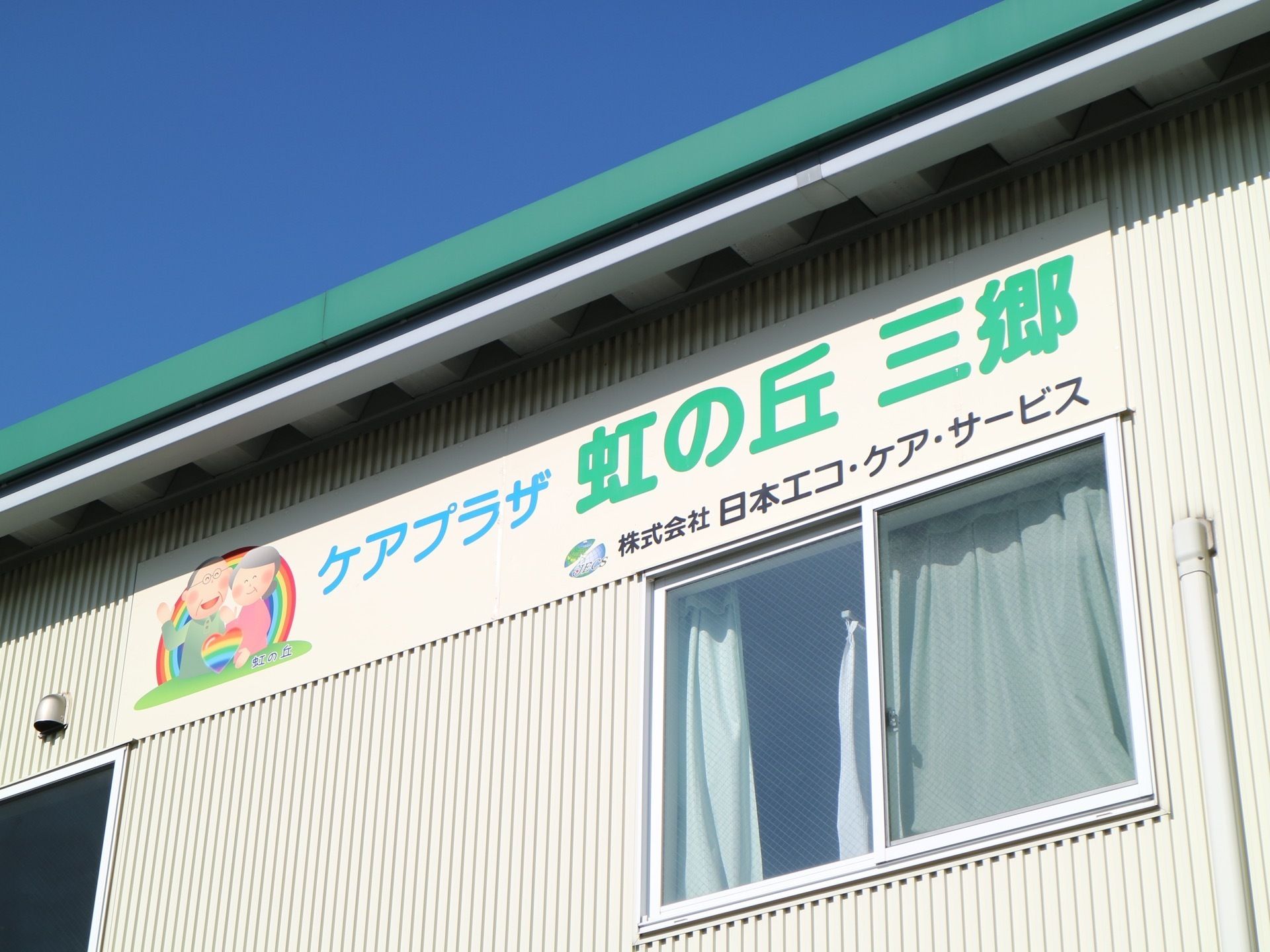 ホワイト調の外壁である。屋根付近に横長の看板が設置されている。ほっこりするイラストのとなりに、施設の名前が書かれている。