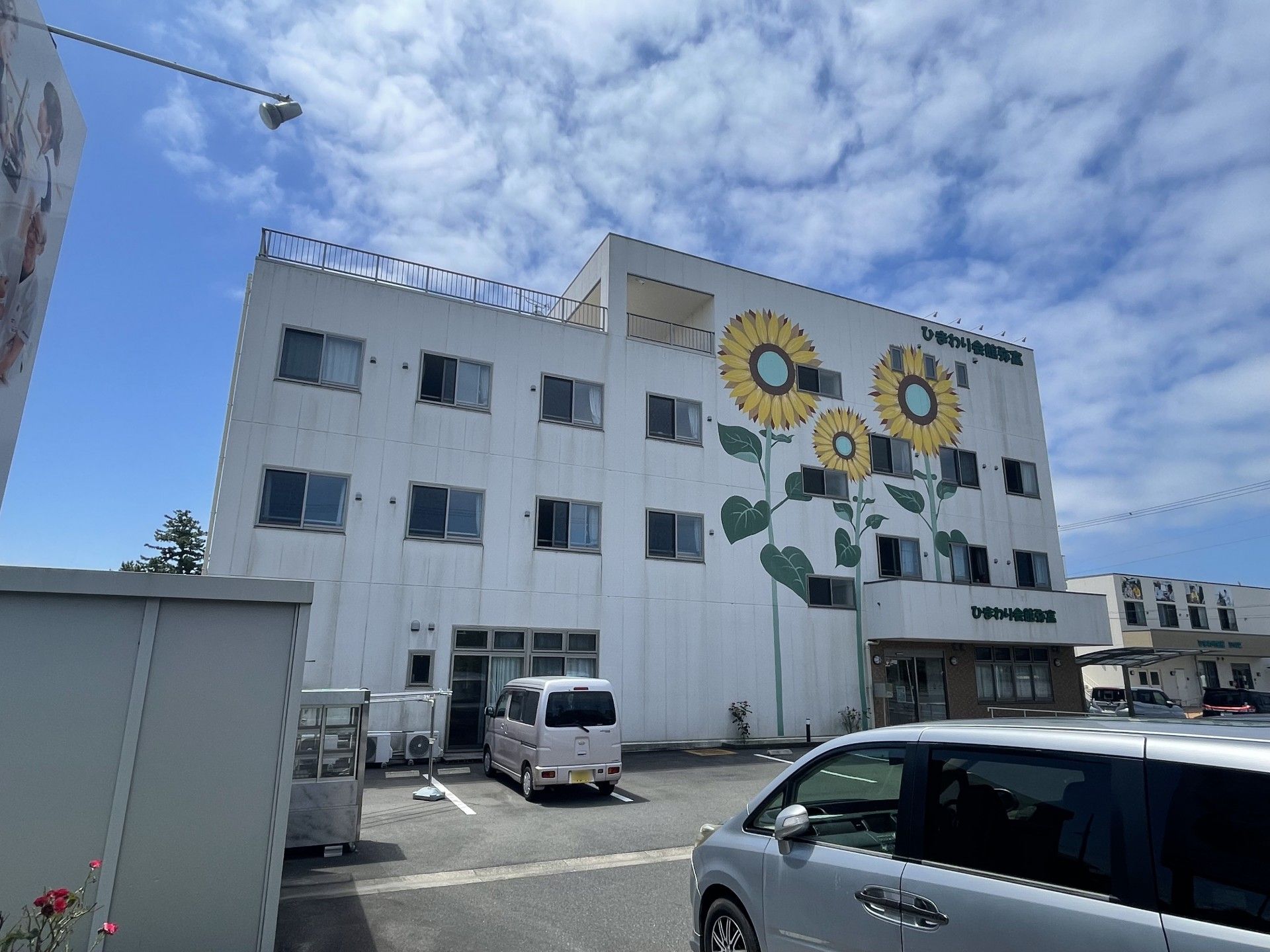 白い外壁の3階建ての建物の玄関には手すり付きのスロープがあり、外壁にはヒマワリの絵が大きく描かれている。