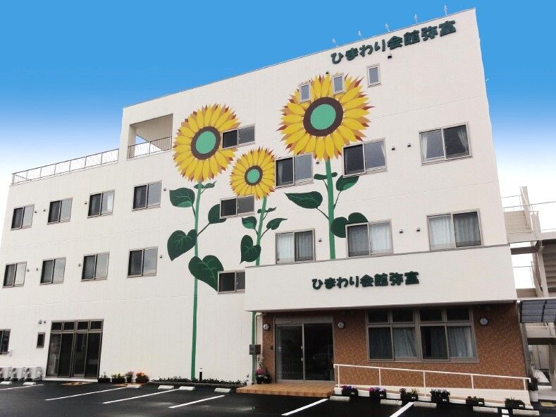 大きなひまわりのイラストが描かれた白い外壁の3階建ての建物の玄関には手すり付きのスロープがあり、駐車場もある。