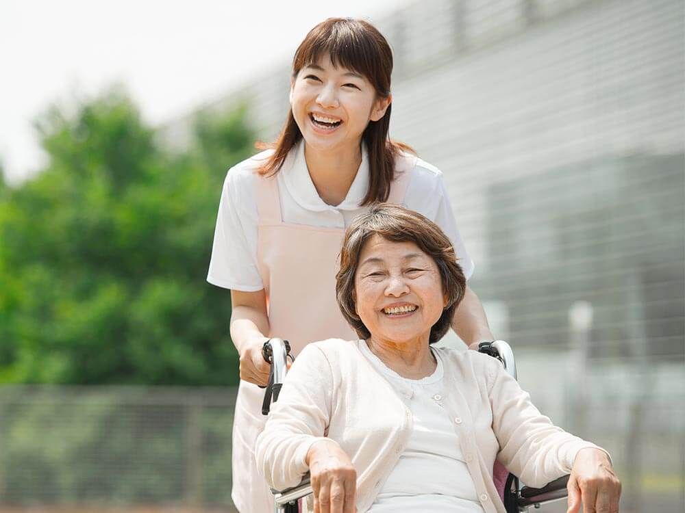 遠くを見ながら笑顔で話している高齢女性と車いすを押す女性スタッフ。散歩の一場面
