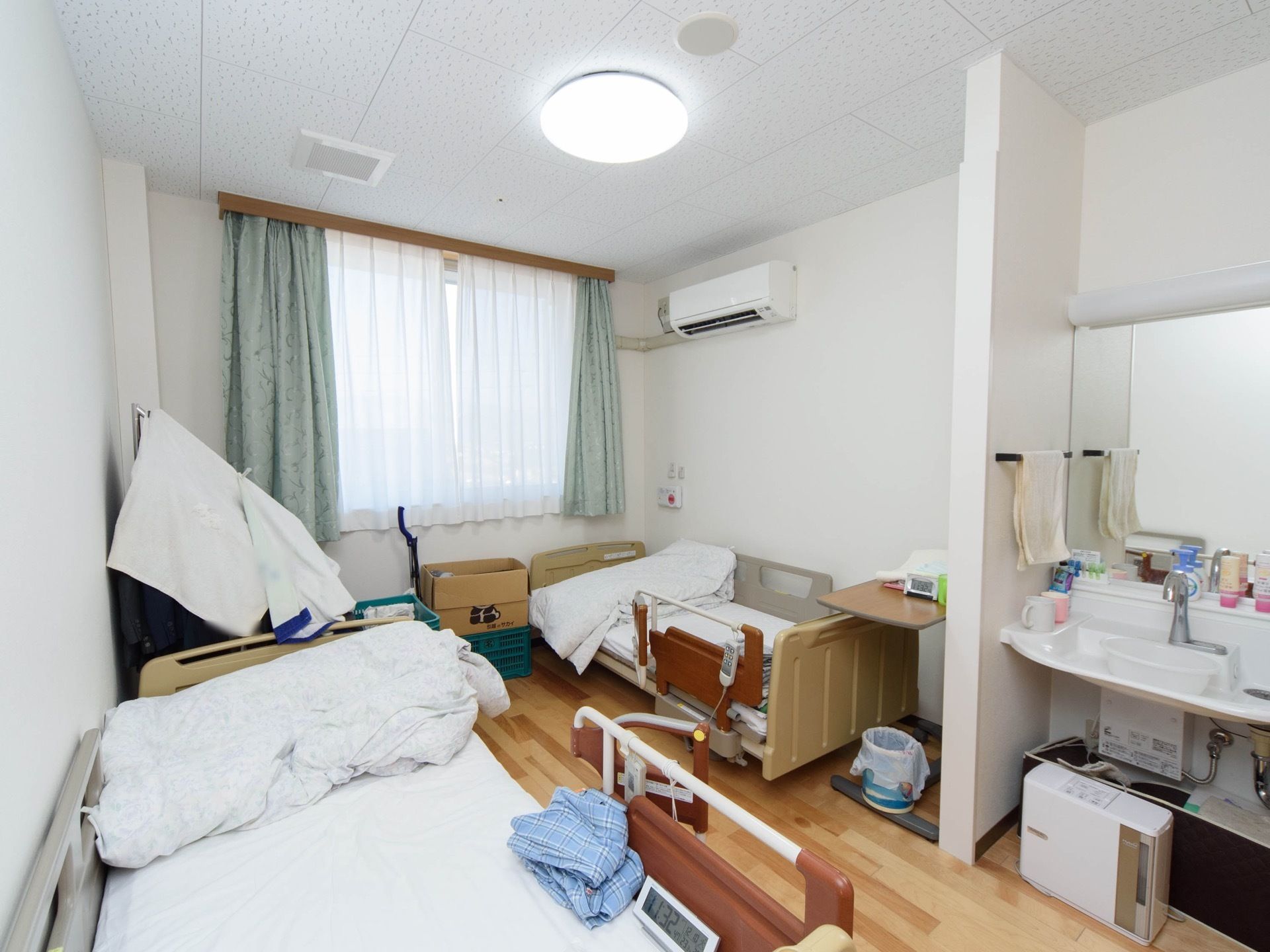 居室にある2台並んだ介護用ベッドに、リモコンとガードが付いている。天井にはエアコン、手前に大きな鏡と洗面台が見える。