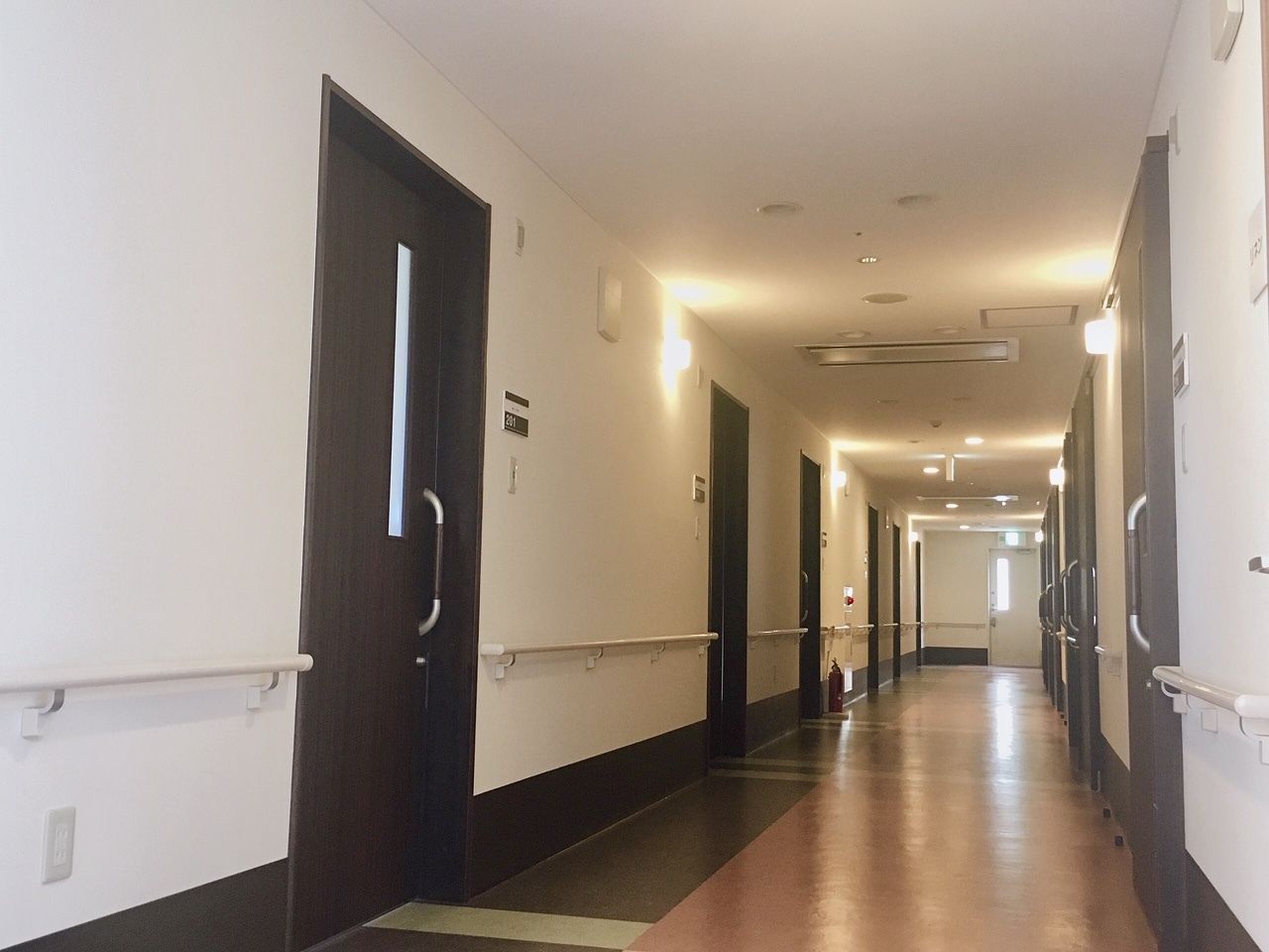 手すりが両サイドについたまっすぐな廊下。居室には引き戸と表札が設置されている。