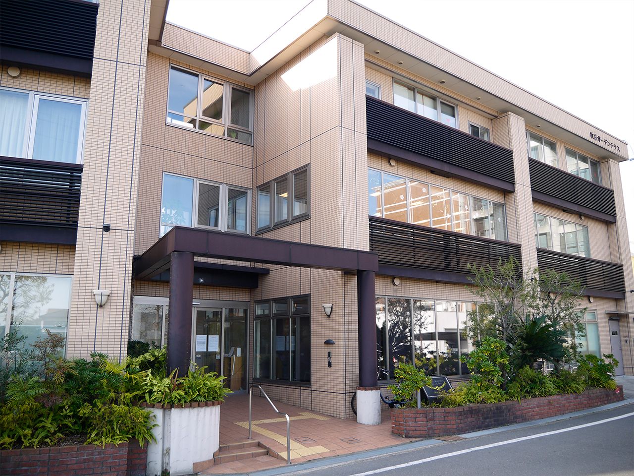 ベージュの３階建ての建物はベランダ付きで、玄関には手すり付きのスロープと階段があり、緑も植えられている。