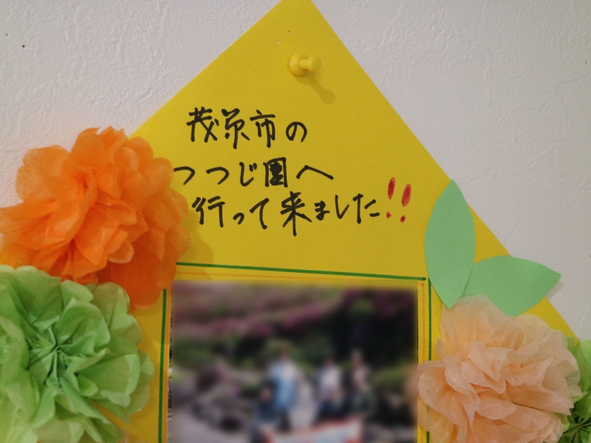 白色の壁に画鋲で貼られている掲示物である。黄色の台紙に紙で作られた花が装飾されており、中央には写真が貼られている。