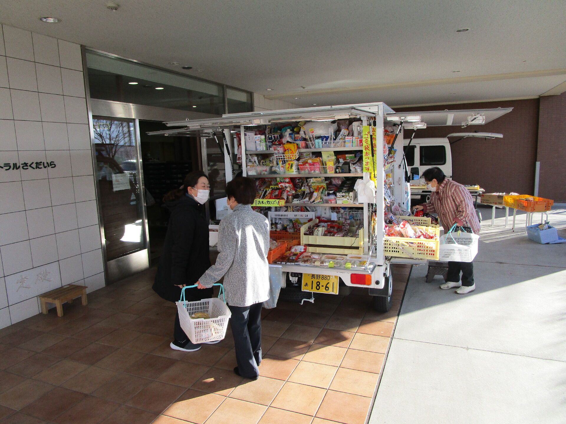 食料品が山ほど詰められた移動式スーパーの車。後ろのドアも横のドアも開く仕組みになっていて、数名の高齢者が買い物かごを手にして買い物をしている。