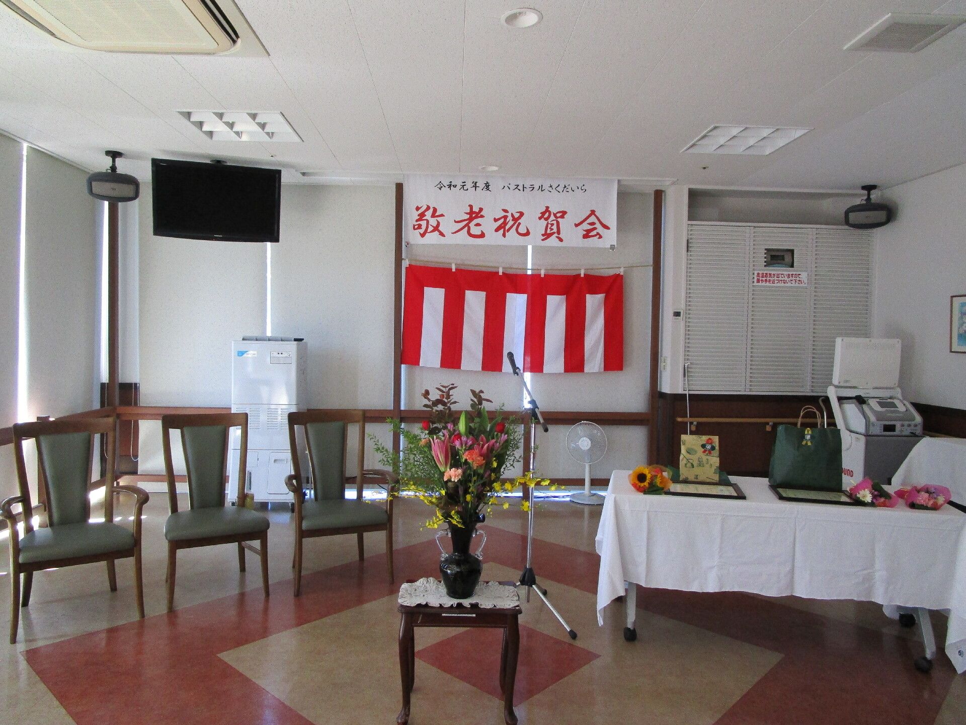 「敬老祝賀会」と書かれた垂れ幕が飾られ、花が飾られたテーブルの後ろにはいすが並べられていて、白いテーブルクロスがかけられた長テーブルもある。