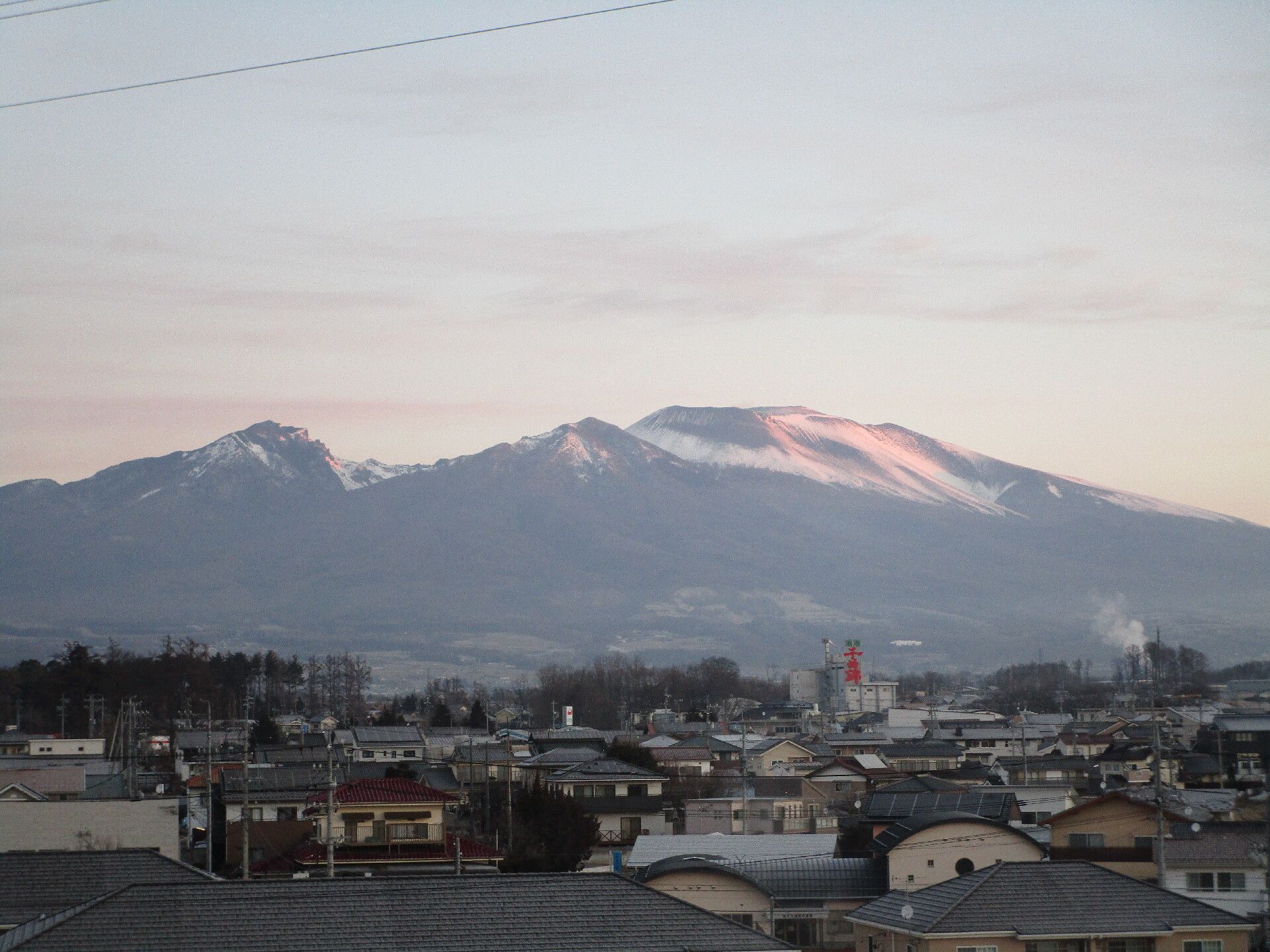 雪がまだ残る山脈が遠くに見え、手前には住宅地が見える。山脈の雪は太陽に照らされてオレンジがかった白になっている。
