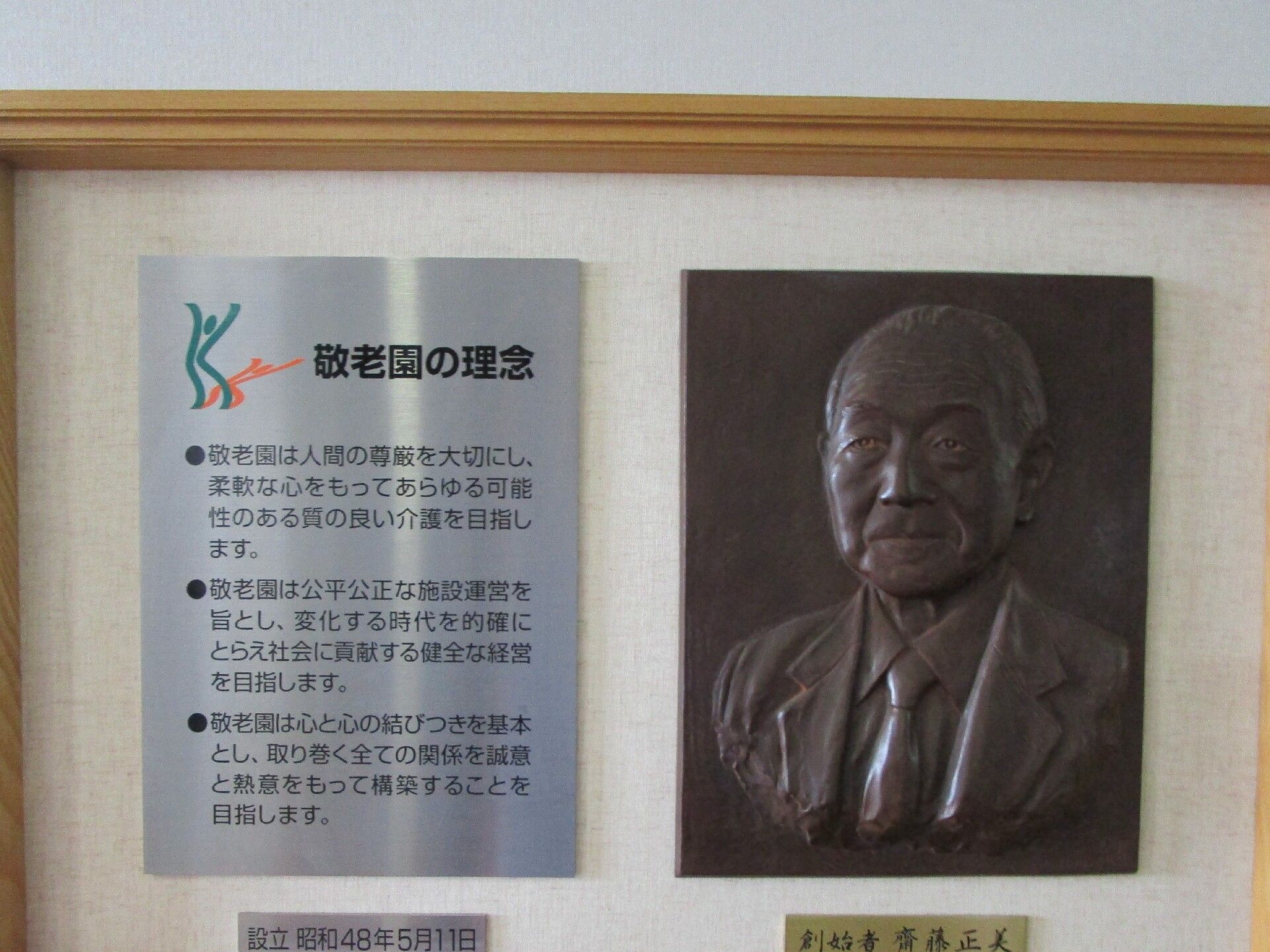 法人の理念である３つの文章が書かれたシルバーのプレートと創設者である男性の姿が彫られた銅版が木製の額の中に飾られている。