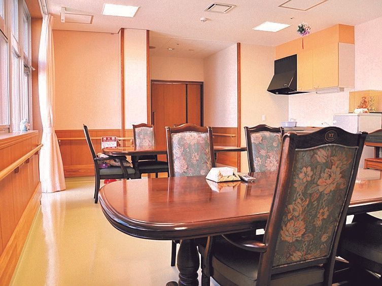 花柄の椅子がセットされた楕円形のテーブルセットが複数並んだ食堂。右の奥には黄色い収納スペースが付いた本格的なキッチンもある。