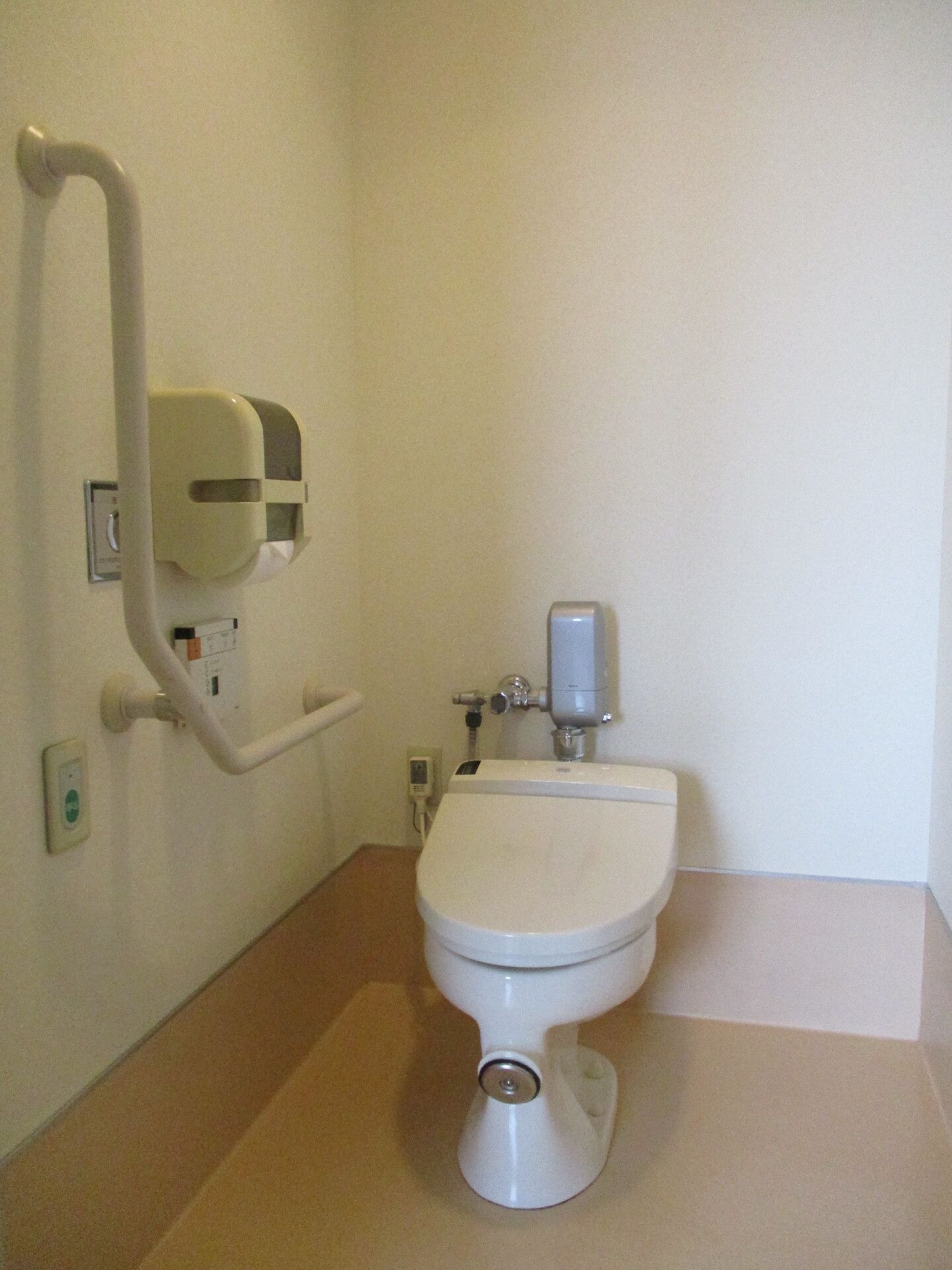 シンプルな白いトイレが付き、壁にはエル字型の手すりやグリーンの呼び出しボタン、トイレットペーパーホルダーなどが付いている。