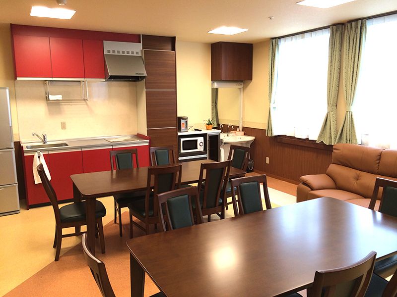 赤いキッチンが奥にあり、手前にはダークブラウンの木製のダイニングテーブルセットが複数置かれ、窓際にはソファも置かれている。