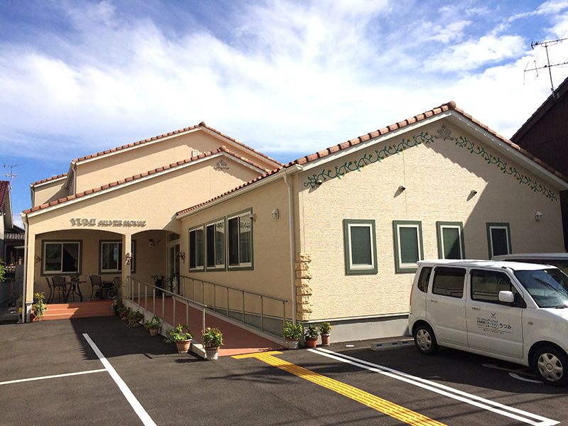 三角屋根が付いた平屋建ての建物。明るい雰囲気の建物で、玄関部分には手すり付きのスロープがあり、外壁にはホーム名が書かれている。