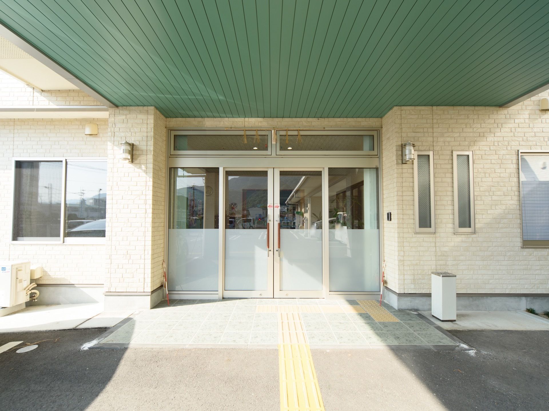 正面から見た施設の玄関。柱付きのひさしが取り付けられた構造になっており、天井は緑色をしている。地面には点字ブロックがある。
