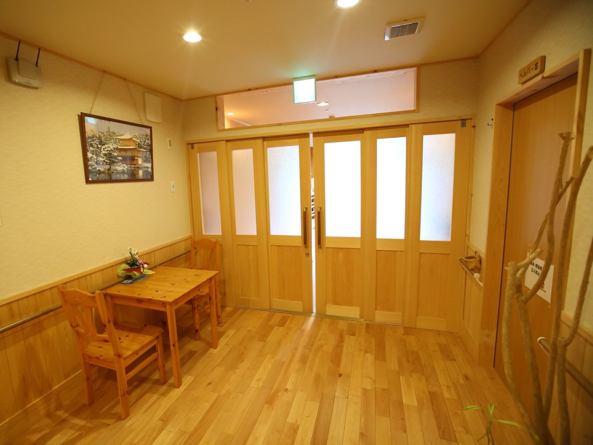 広々とした温かい雰囲気の廊下である。右にヘルパー室が設けられており、左の壁際に２人掛けテーブルが置かれている。