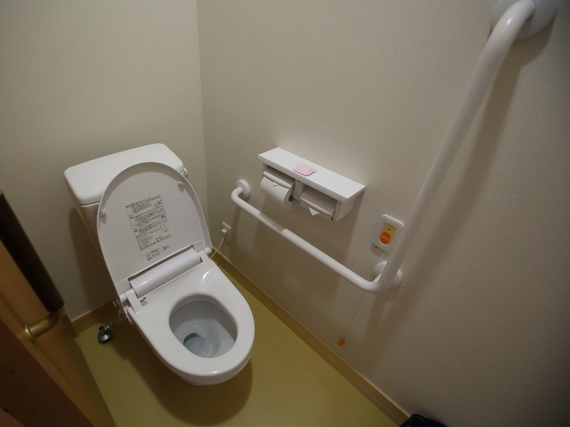 居室のトイレである。L型手すりと呼び出しボタンが壁に設置されている。扉には、引き戸が採用されている。