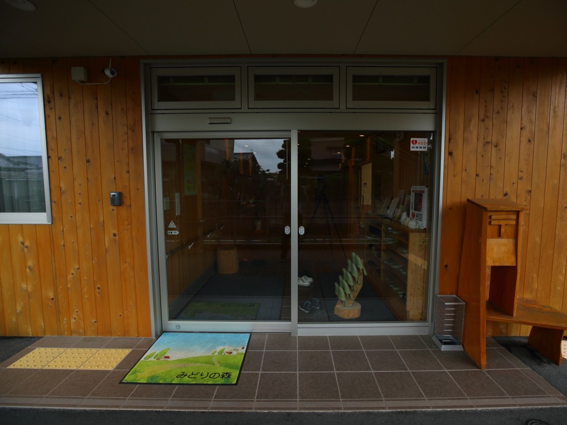 大きな屋根付きの広いエントランスである。壁には、インターホンや防犯カメラが設置されている。木製の郵便受けが置かれている。
