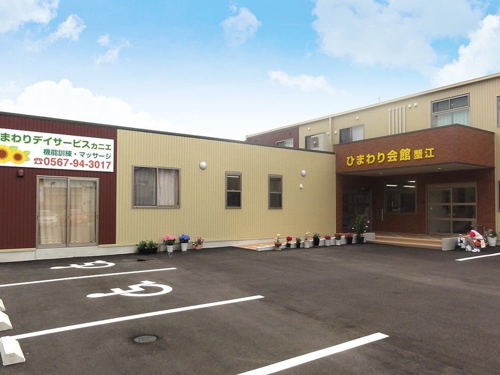 ブラウン系の建物が建つ敷地内には駐車場があり、玄関の周りには花が植えられたプランターがたくさん置かれている。