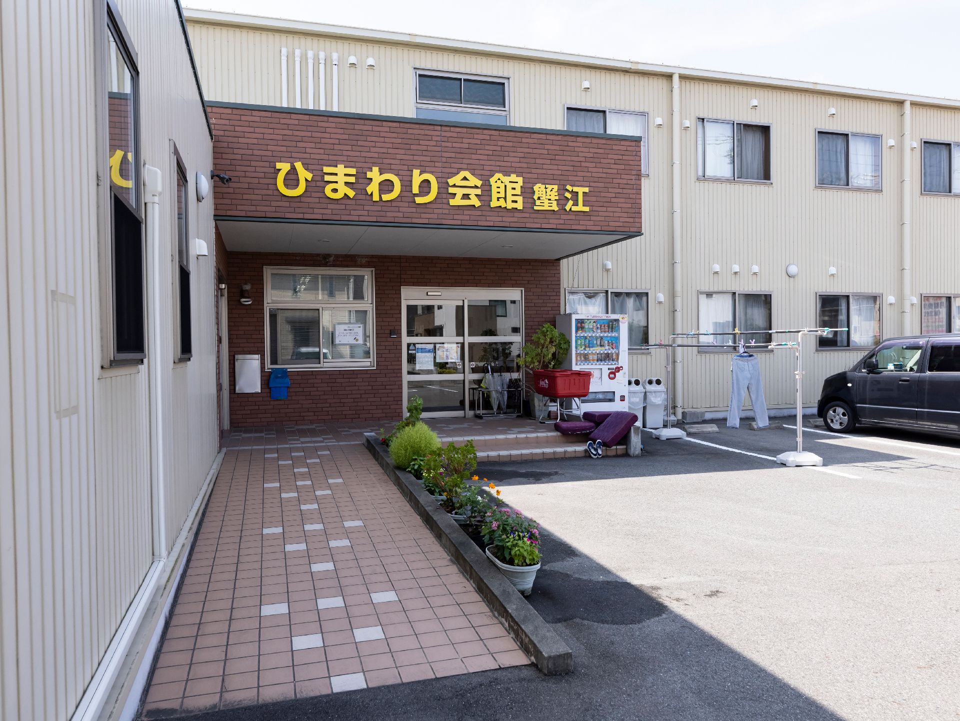 白い花がたくさん咲く道を楽しそうに散歩している車いすの高齢女性と付き添いの高齢男性。女性スタッフも近くにいる。