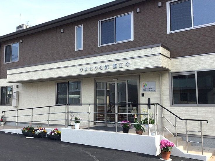ダークブラウンとアイボリーの2階建ての建物は玄関先に階段と手すり付きのスロープが付いていて、花も多い。