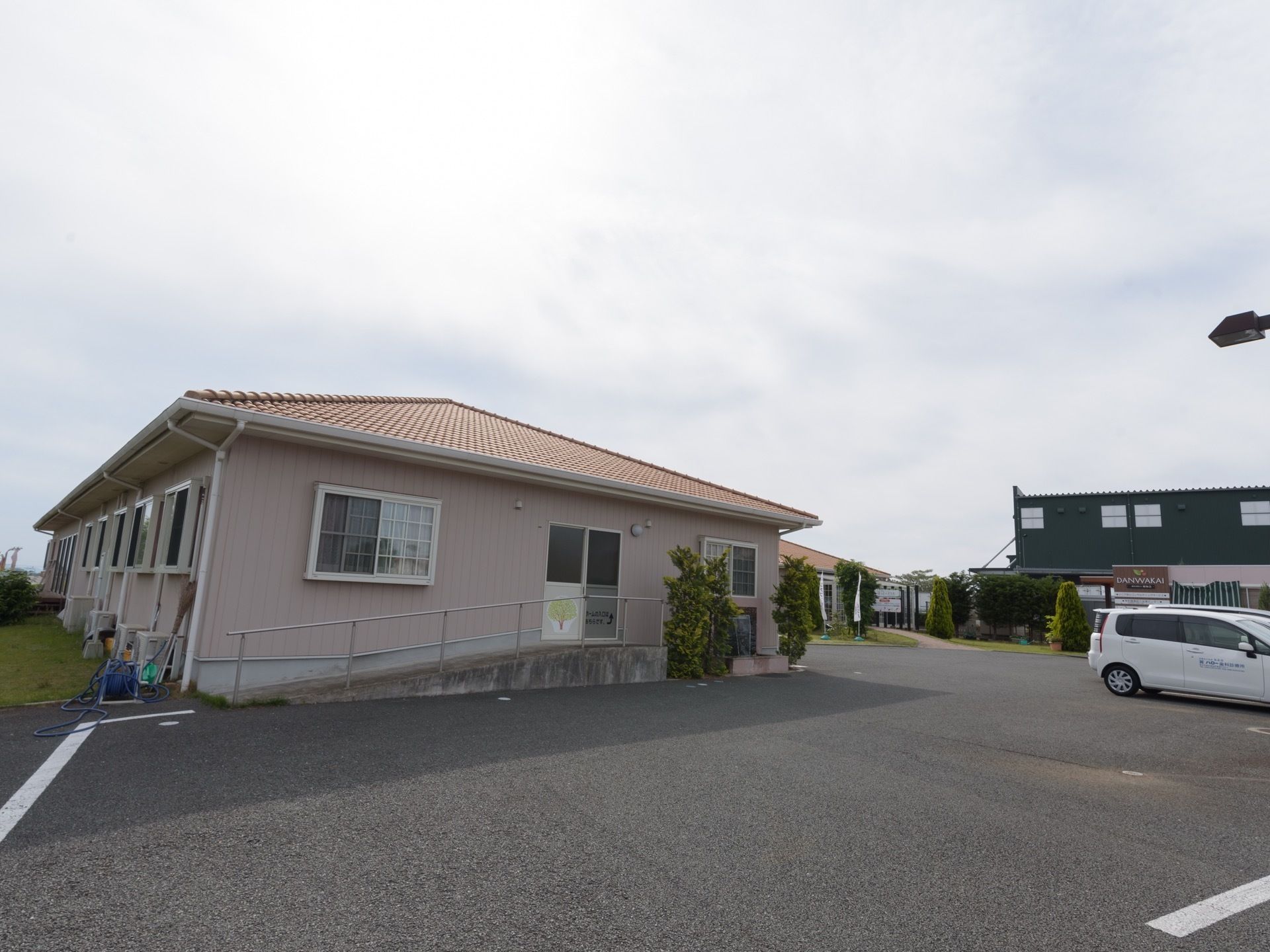 舗装された敷地に、平屋建ての住宅が建っている。周りは芝生に囲まれていて、建物の前は広い駐車スペースになっている。