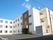 広い駐車場が付いた白を基調とした建物は3階建てで建物の周りには草木が植えられている。