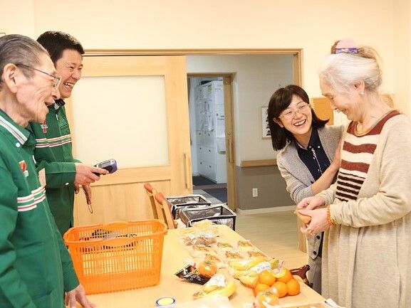 セブンイレブンのユニフォームを着た男性スタッフが2名、高齢女性の接客をホーム内でしている。