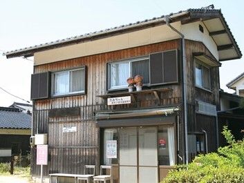 2階建ての建物は瓦屋根が付きアットホームな雰囲気。玄関先にはいすが2脚並び、ベンチも置かれている。周りには住宅が建っている。