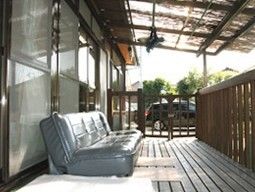 グレーのソファが置かれたウッドデッキのテラスは日当たりがよく、屋根も付いている。木製の柵で囲まれていて、柵の向こう側には駐車場もある。