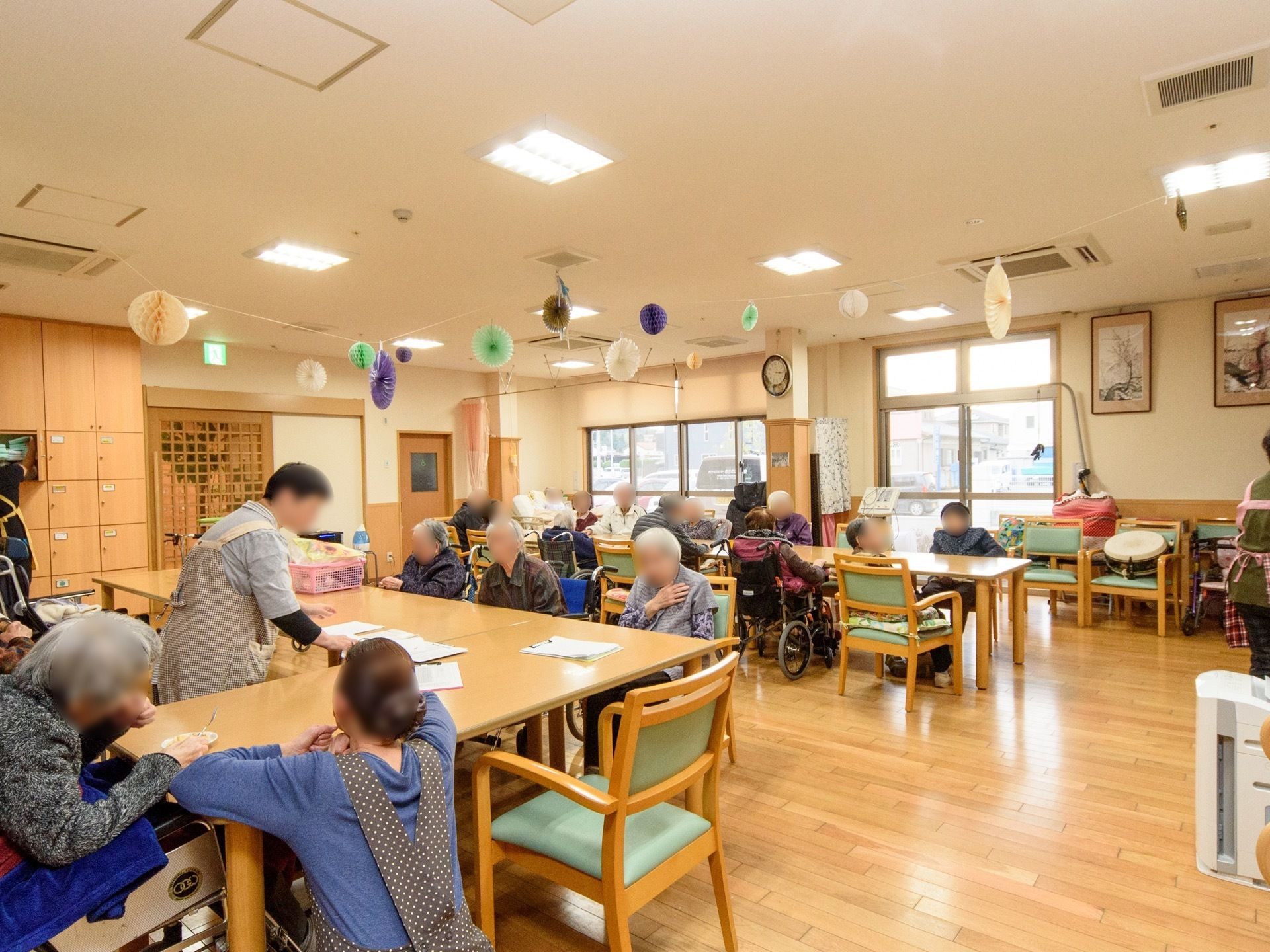 共有スペースに多くの入居者様が集まっている。エプロンをした女性スタッフが３名、入居者様のお世話をしたり、作業をしたりしている。