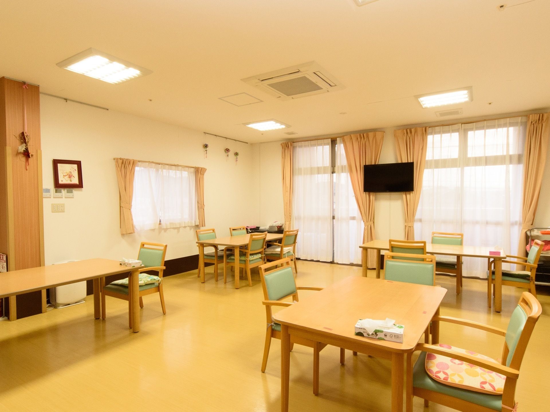 食堂に掃き出し窓や腰高の窓がある。窓の間の壁の上に方にテレビが掛けられている。天井埋め込み式のエアコンが設置されている。