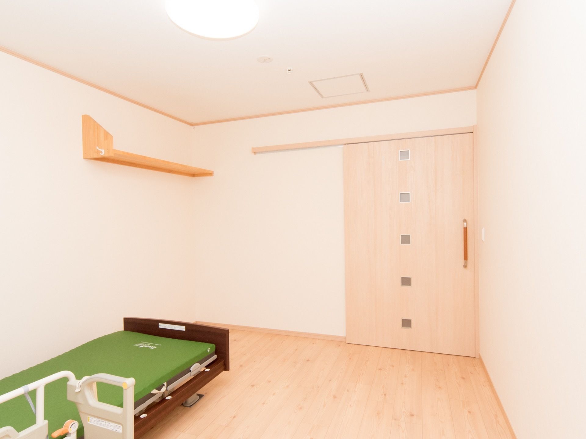 白みがある木調の床と建具が明るい印象を与えている。枠が木製の介護ベッドが完備されている。壁には棚がある。