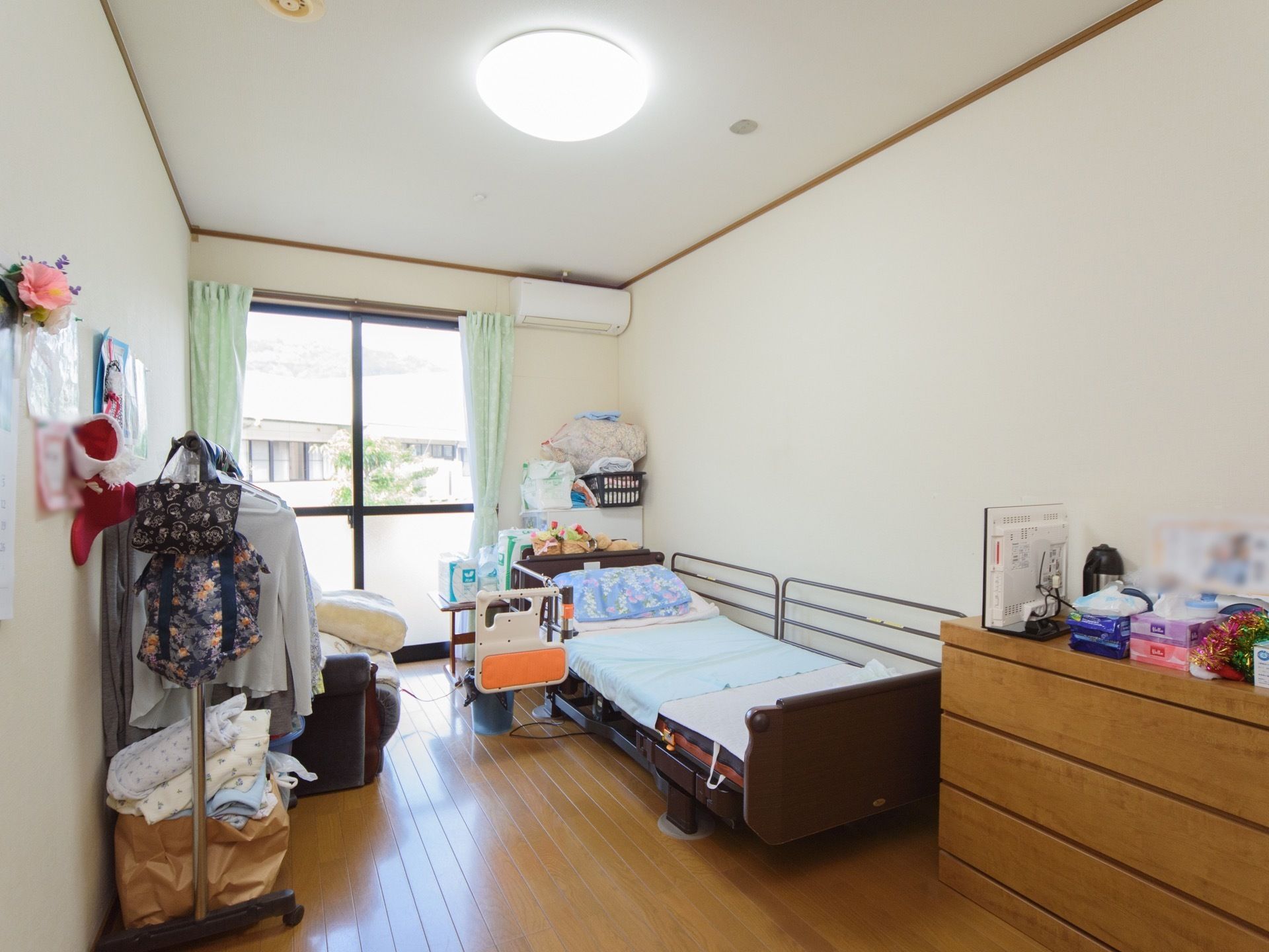 居室のベッドは、柵で塞がれている側と、開閉できる柵が付いた側とがある。窓には緑色のカーテンを付け、右横の壁にエアコンがある。