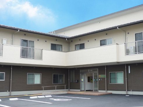 2階建ての建物は1階部分がブラウン、2階部分が白でベランダが付き、玄関には手すりがある。