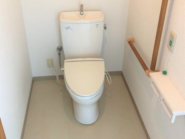 エル字型の木製の手すりが設置されたトイレ。便座はシンプルなデザインで、壁には呼び出しボタンもある。
