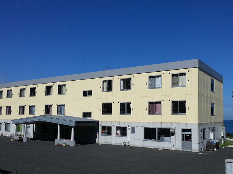 3階建ての建物の１階部分はライトグレー、２階と３階はベージュの外壁で、玄関には車寄せが付いている。