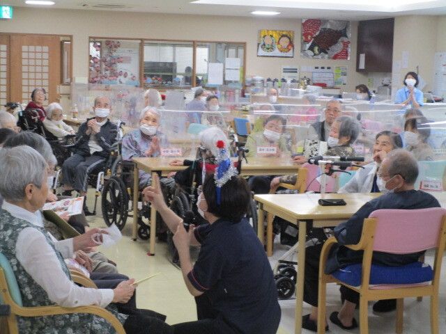 ポロシャツを着た女性スタッフが司会を務めるイベント。テーブルや壁際の椅子には多くの高齢者がいる。