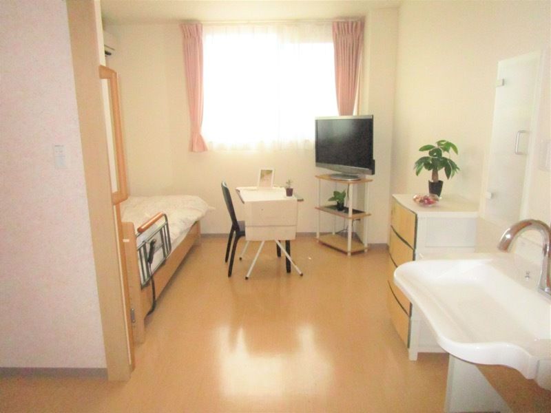 レースカーテンとカーテンが付いた大きな窓があるフローリングの部屋には寝具付きのベッドやテレビがある。