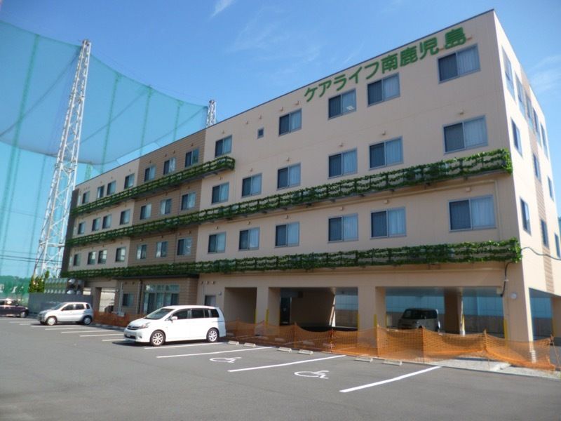 ベージュ系の4階建ての建物が建っている敷地内には駐車場があり、建物には緑の文字が書かれている。